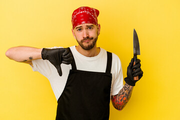 Young tattooed batcher caucasian man holding a knife isolated on yellow background showing a dislike gesture, thumbs down. Disagreement concept.