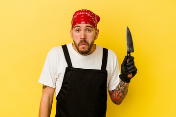 Young tattooed batcher caucasian man holding a knife isolated on yellow background shrugs shoulders and open eyes confused.