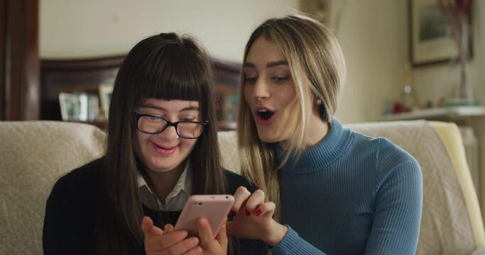 Cinematic shot of happy sister and teen girl with down syndrome using together smartphone for family entertainment at home. Concept of technology, connection, handicapped, disability, online, fun.
