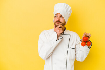Young caucasian chef man holding tomatoes isolated on yellow background looking sideways with doubtful and skeptical expression.