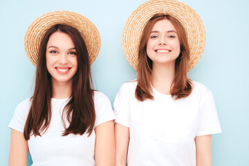 Two young beautiful smiling hipster female in trendy same summer white t-shirt and jeans clothes. Sexy carefree women posing near light blue wall in studio. Cheerful and positive models in hats