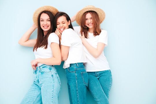 Three young beautiful smiling hipster female in trendy same summer white t-shirt and jeans clothes. Sexy carefree women posing near light blue wall in studio. Cheerful and positive models in hats
