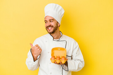 Young caucasian chef man holding eggs isolated on yellow background points with thumb finger away, laughing and carefree.
