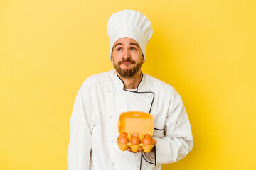 Young caucasian chef man holding eggs isolated on yellow background dreaming of achieving goals and purposes