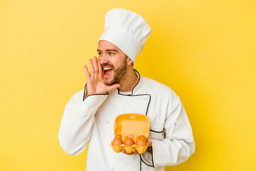 Young caucasian chef man holding eggs isolated on yellow background shouting and holding palm near opened mouth.