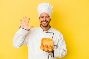 Young caucasian chef man holding eggs isolated on yellow background smiling cheerful showing number five with fingers.