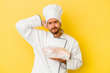 Young caucasian chef man holding chicken isolated on yellow background touching back of head, thinking and making a choice.