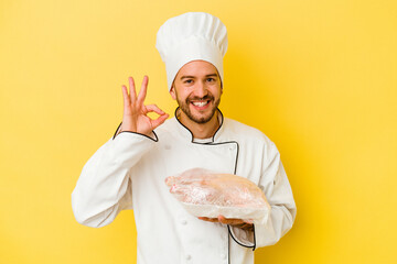 Young caucasian chef man holding chicken isolated on yellow background cheerful and confident showing ok gesture.