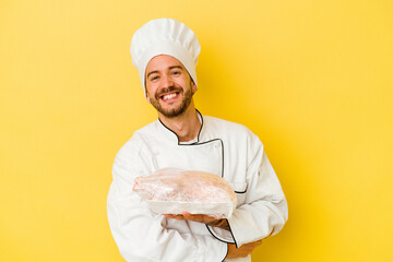 Young caucasian chef man holding chicken isolated on yellow background laughing and having fun.