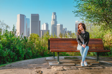 Stunning brunette woman in a denim jacket sits on a park bench with a view of Downtown skyscrapers...