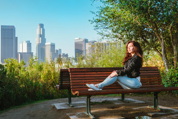 Stunning brunette woman in a denim jacket sits on a park bench with a view of Downtown skyscrapers in Los Angeles