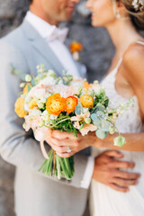 The bride and groom stand hugging at a brick wall in the old city and hold a wedding bouquet with orange flowers, close-up 