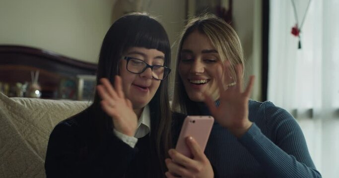Cinematic shot of happy sister and teen girl with down making video call together with smartphone to friends or family at home. Concept of technology, handicapped, disability, media, love, friendship