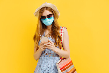 Young woman in medical mask, in summer dress and hat uses mobile phone isolated over yellow background