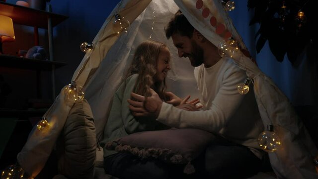 Bearded father tickling and hugging his teen daughter while they sitting in decorative makeshift tent. Dad and girl laughing and having fun together. Concept of happy family time.