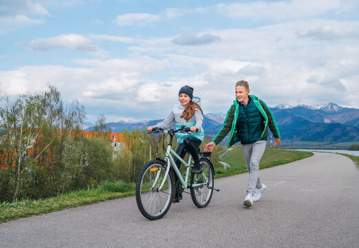 Smiling kids on a bicycle path with snowy mountains background. Brother helping to sister and teaching doing first steps in riding. Happy childhood concept image.