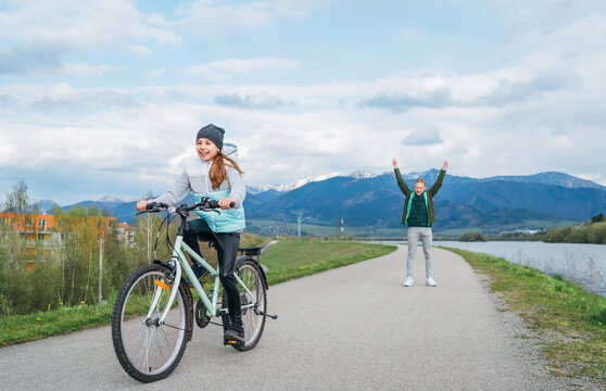 Smiling kids on a bicycle asphalt path with snowy mountains background. Brother helping to sister. He rose arms up after a successful riding start. Happy childhood concept image.