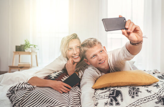 Cheerful young adults сouple in pajamas taking a selfie photo using a modern smartphone as they lazy relaxing lying in a cozy bed in the sunny bedroom. Couples relations concept image.