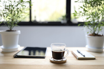 Black coffee in transparent glass with tablet and notebook with chili plant pot on wooden table