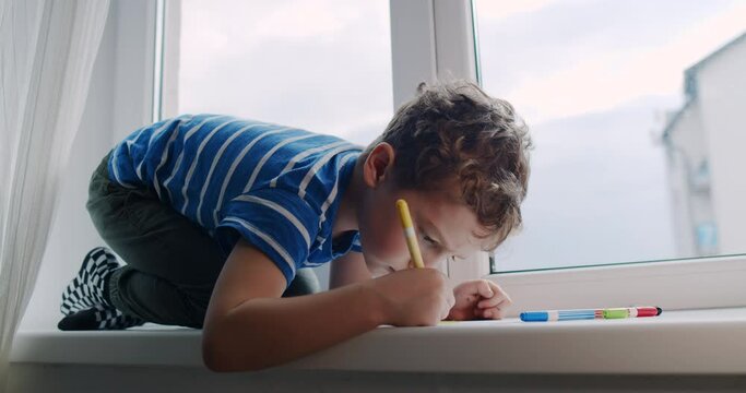 Little boy with curly hair draws on the window at home with crayons. 
