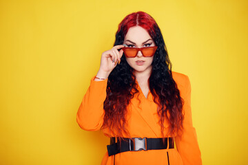 Beautiful girl on a yellow background. A brunette in an orange dress and long hair is posing, smiling, having fun. Woman in sunglasses. Joy and emotion. Red hair roots place for text