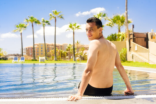 A man sits by the pool with his feet in the water. Sunny day, a man enjoys relaxation. Travel. A man sits on a background of palm trees and a house. A man in a hotel with a pool.