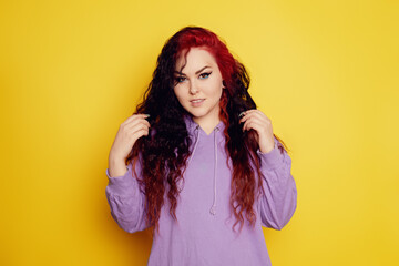 Woman in a purple sweater on a yellow background posing. A beautiful brunette with long curly hair and red roots. Eyeliner and trendy makeup. Model looking at the camera