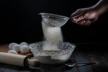 Sifting flour into a cristal bowl with eggs on the side and on a table, with a black background, bakery, or cooking concept dark food or low key light photography