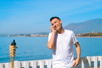 A man speaks on the phone against the backdrop of the beach and ocean, sea. Guy in a white shirt on vacation. Freelancer calls and smiles, roaming and chatting. Travel