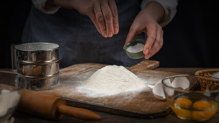Women's hands, flour and dough. A woman in an apron prepares dough for homemade baking, a rustic home cozy atmosphere, a dark background with unusual lighting.