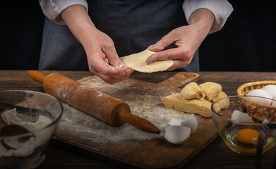 Women's hands, flour and dough. A woman in an apron cooking dough for homemade baking, a rustic home cozy atmosphere, a dark background with unusual lighting.