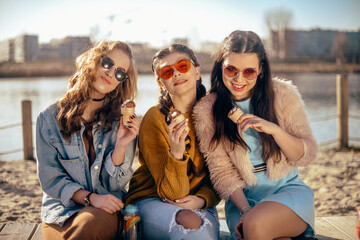 Three girls sit on the beach , near the river, talk, gossip, joke, laugh, smile at womans day. Girls on a hot day eat ice cream. Beauty model wearing sunglasses at summer day. Girl with hairslyle