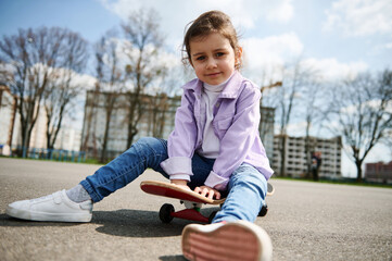 Cute female child posing to the camera while having rest sitting on a wooden skateboard on the playground