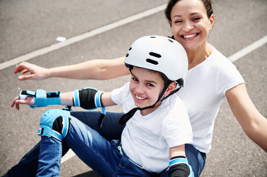 Happy mom and adorable son in protective helmet and equipment enjoying time together sitting on a skateboard