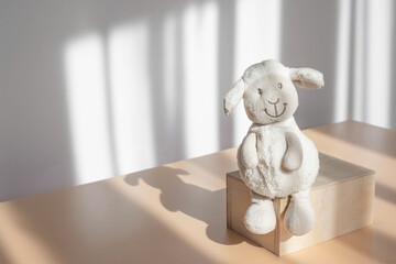 Lonely children's toy plush white lamb sitting on wooden stand on background of white wall with shadows. Stuffed toy of animal for baby. Copy space