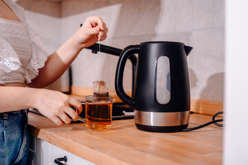 Fototapeta na wymiar A black kettle is on the kitchen table. A woman's hand holds a teapot and makes tea. Tea bag in a mug with water, tea leaves.