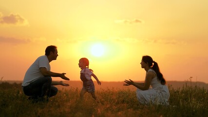 Fototapeta na wymiar Happy family walks in park at sunset. Mom, dad and baby. Little daughter goes from mom to dad, hugs and kisses her parents in rays of a warm sun. Healthy family plays in field. Happy family concept