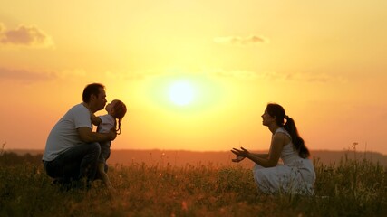 Fototapeta na wymiar Happy family walks in park at sunset. Mom, dad and baby. Little daughter goes from mom to dad, hugs and kisses her parents in rays of a warm sun. Healthy family plays in field. Happy family concept