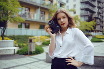 Fototapeta premium A beautiful girl looks in the viewfinder in the camera, and takes pictures. The blonde learns to photograph and earn money on the photo. A girl photographer walks around the city and takes pictures