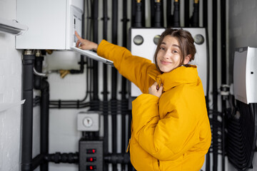 Young woman feeling cold, wearing jacket in the boiler room. Housewife adjusts temperature on the gas boiler at home