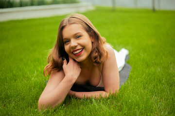 Naklejka na ściany i meble A teenager girl lies on the grass in a beautiful white dress, rests, enjoys nature. Girl with short blond hair. Beautiful and healthy skin and youth smile