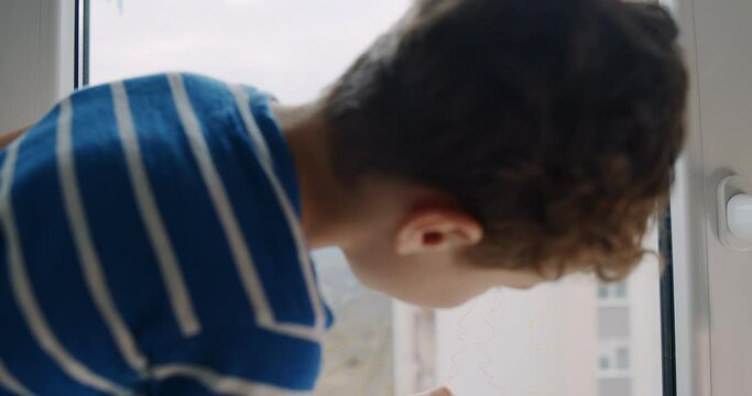 Little boy with curly hair draws on the window at home with crayons. 