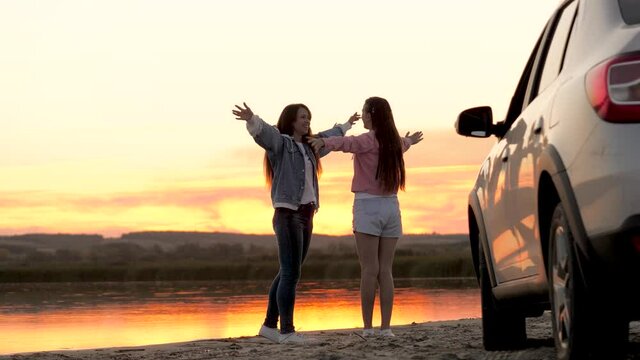 Free women travelers stand next to car on beach enjoying sunset in park, jumping and clapping rejoicing. Girls drivers stopped at campsite. Girlfriends are enjoying trip in car. Vacation, adventure