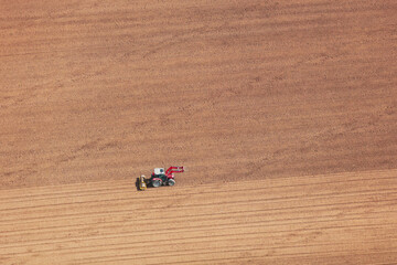 An aerial view taken from a helicopter of a red tractor crossing an empty farm field in Britain. The isolated vehicle is pulling a field roller. The ploughed land makes an abstract striped pattern.