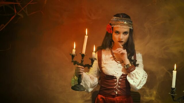 Fantasy woman gypsy in red vintage dress. Girl witch fortune teller holds candlestick burning candles. Room in smoke. Spiritual mystical seance Shows finger gesture to be silent do not make noise.