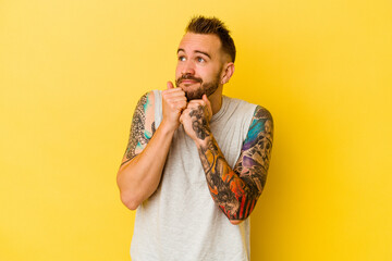 Young tattooed caucasian man isolated on yellow background keeps hands under chin, is looking happily aside.
