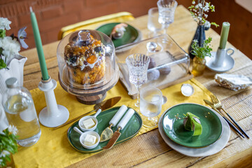 Happy Easter concept. Beautiful spring table settings, green plate with egg, panettone on cake stand, green candles, and branch of apple flowers in vase. Holiday tablescape. 