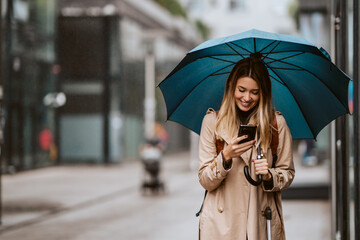 Beautiful girl with an umbrella standing in the rain typing a message. - obrazy, fototapety, plakaty