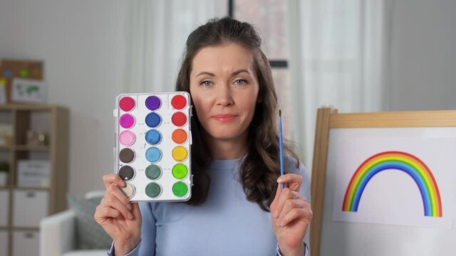 distant education, primary school and teaching concept - female teacher with colors and picture of rainbow having online class of arts at home