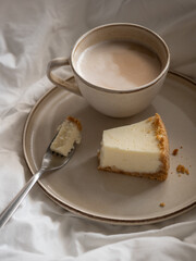 Morning cup of coffee and classic cheesecake on a beige ceramic plate on a soft white bed in the morning light